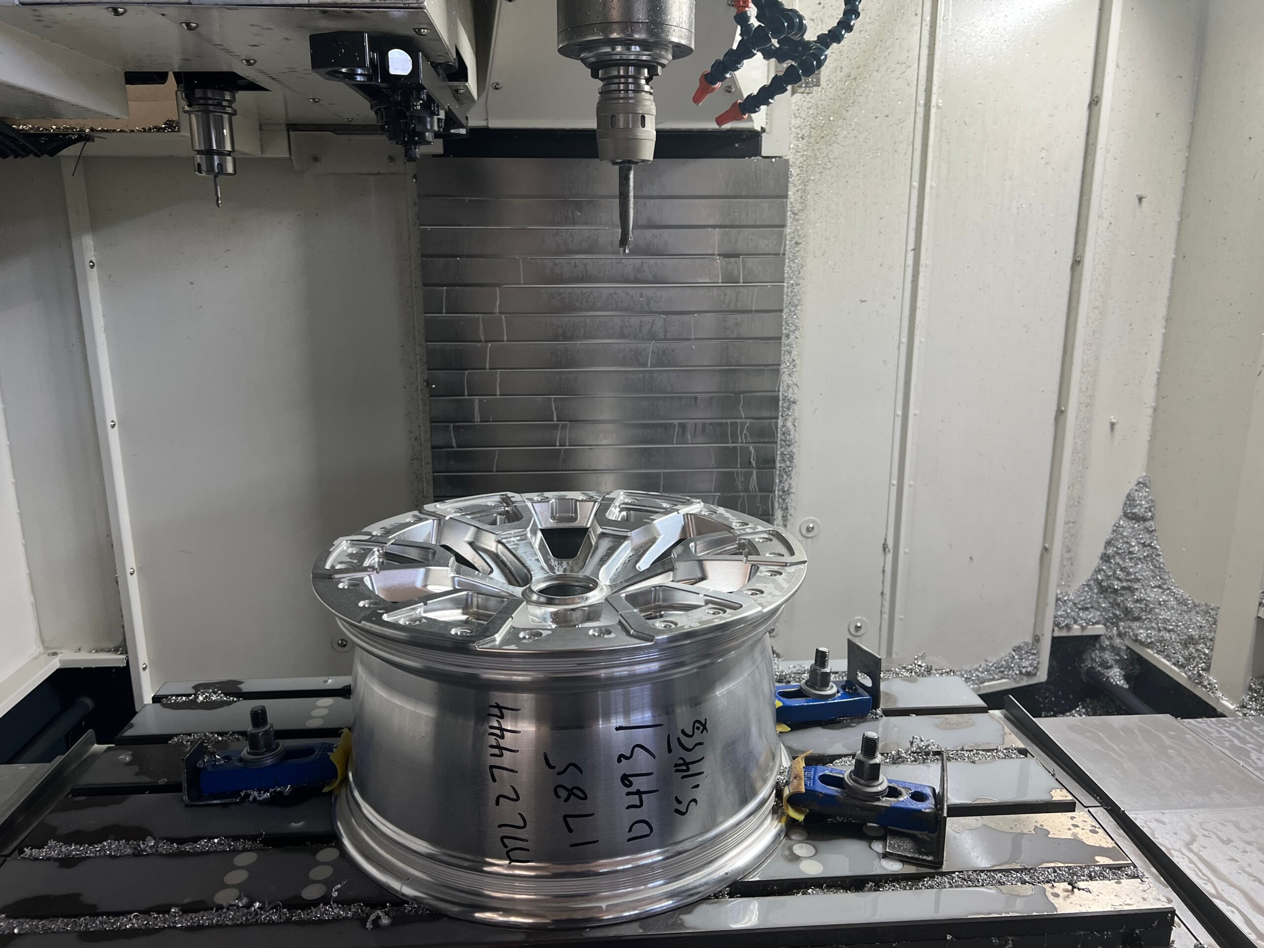 Bare forged aluminium wheel clamped in a CNC machining centre at the FLEXIFORGED factory in Chongqing, with aluminium chips on the machine bed